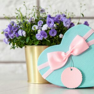 Heart-shaped gift box with bow beside purple flowers, perfect for gifting occasions.