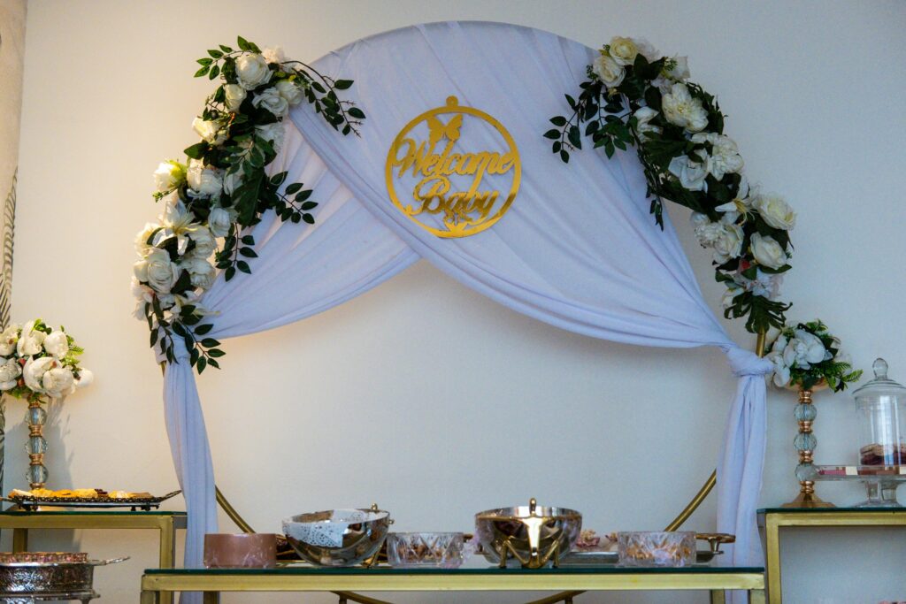 Sophisticated baby shower setup featuring a floral arch with gold 'Welcome Baby' sign and elegant table settings.