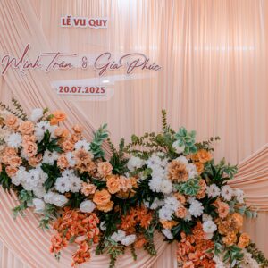 A beautiful wedding floral arrangement with peach and white flowers set against elegant draped fabric featuring names and date.