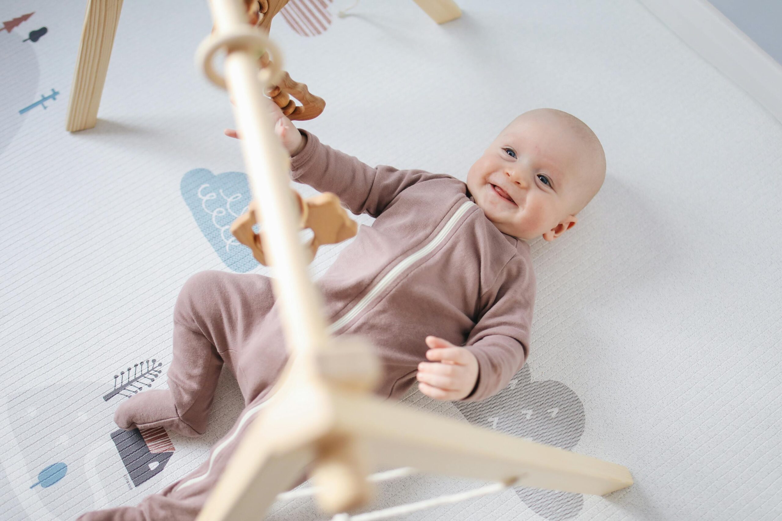 pexels photo 3875215 3875215 A joyful baby lying on a soft mat, playing with wooden toys, smiling brightly.