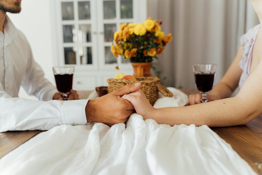 A couple enjoying a romantic dinner indoors, holding hands over a table set with wine and flowers.