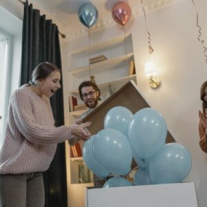 Friends joyfully celebrate a gender reveal with blue balloons indoors.