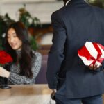 A man in a suit holds a gift box behind his back, preparing to surprise a woman with roses in a cafe.