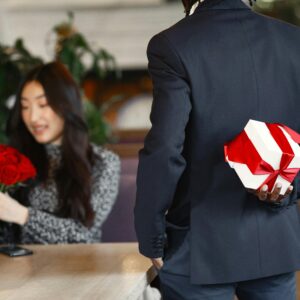 A man in a suit holds a gift box behind his back, preparing to surprise a woman with roses in a cafe.