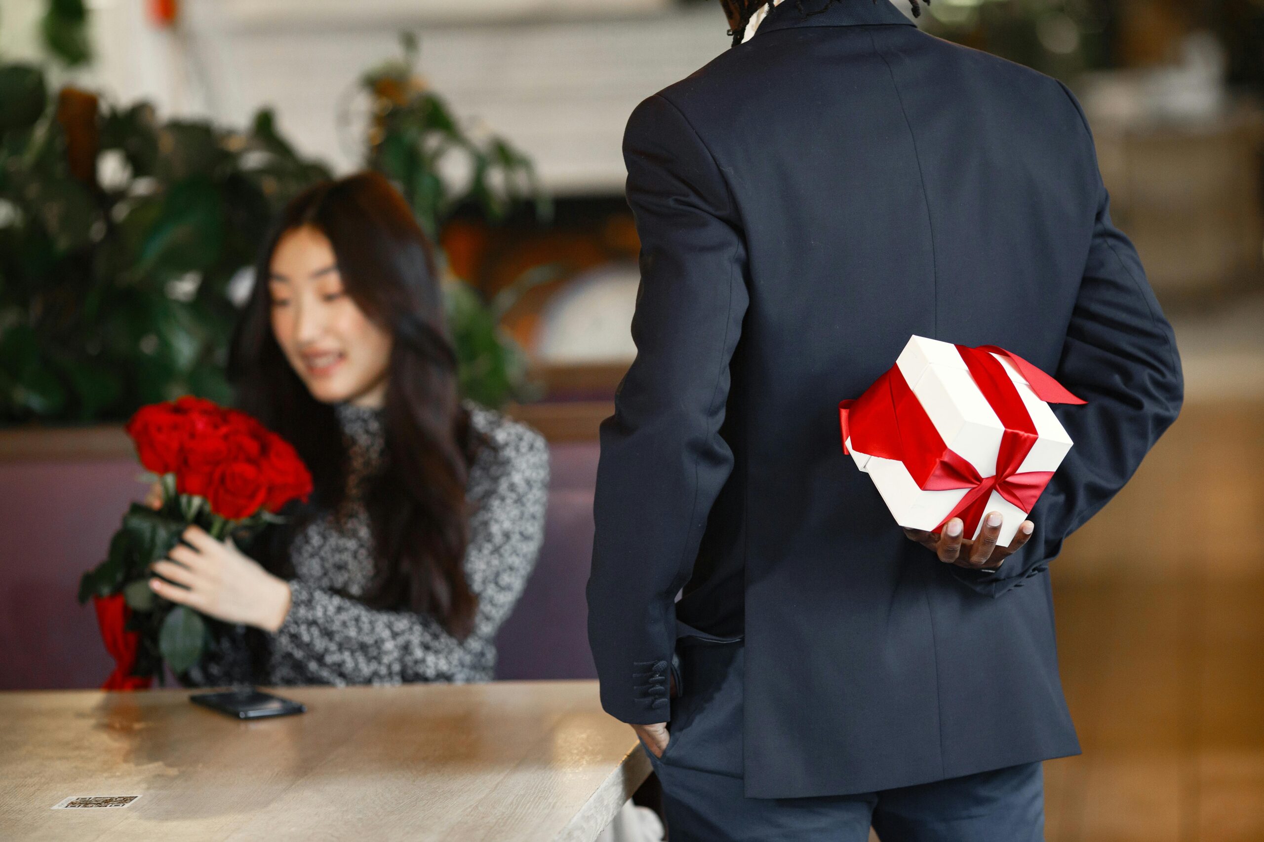 pexels photo 6699481 6699481 A man in a suit holds a gift box behind his back, preparing to surprise a woman with roses in a cafe.