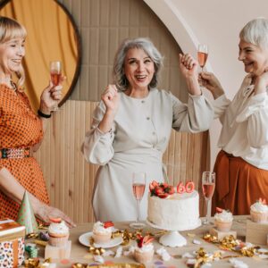 Three senior women celebrating a birthday with cake and champagne indoors.