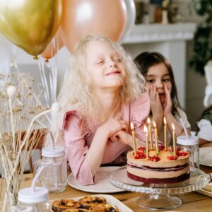 A happy girl makes a wish with lighted candles on her birthday cake, surrounded by friends and balloons.