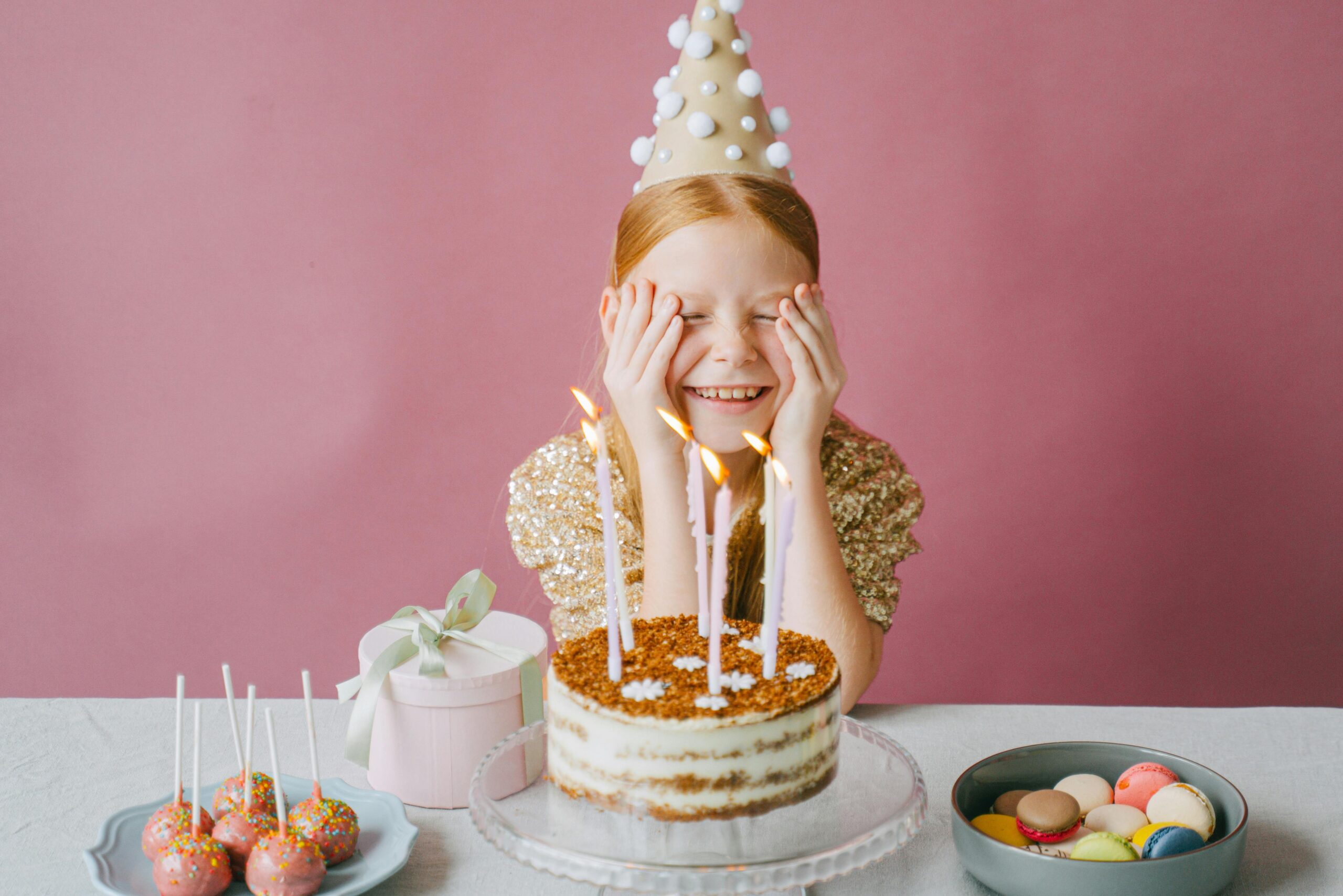 pexels photo 9628330 9628330 A young girl celebrates her birthday with cake and lollipops, wearing a party hat and gold dress.
